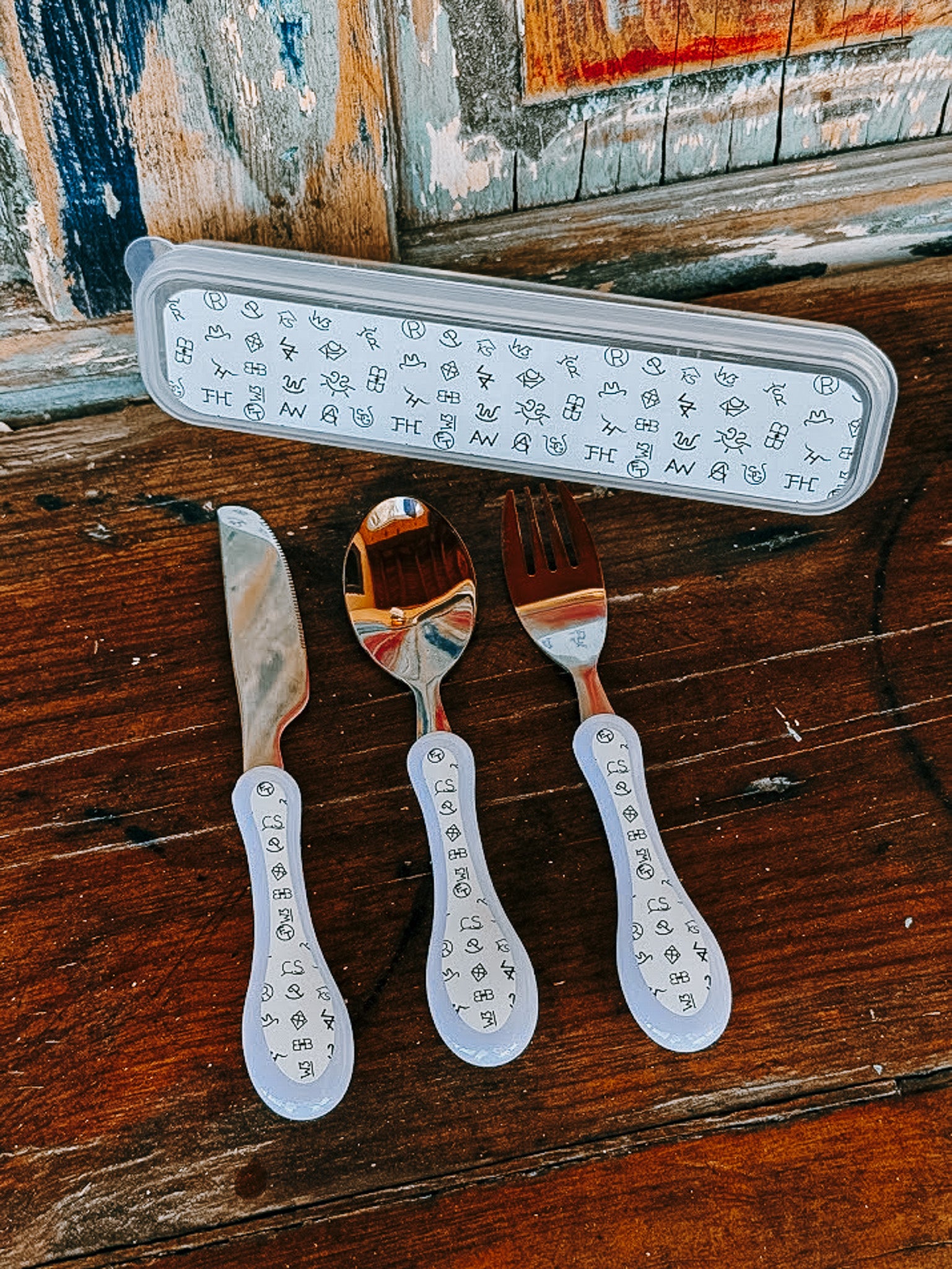 Three personalized stainless steel cutlery pieces with white handles printed with black monogram letters, displayed on a rustic wooden table next to their matching storage case.