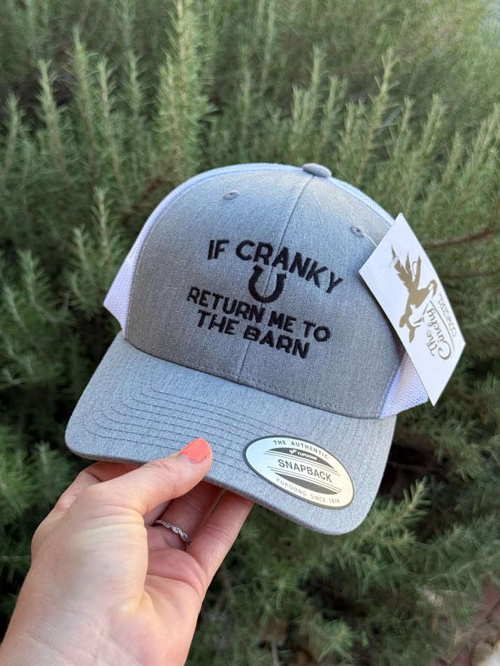 A light gray heather trucker hat held by a hand, featuring black embroidered text that reads 'IF CRANKY RETURN ME TO THE BARN' with a horseshoe graphic. The hat has a white hangtag and a silver Snapback sticker on the brim, set against a background of green foliage.
