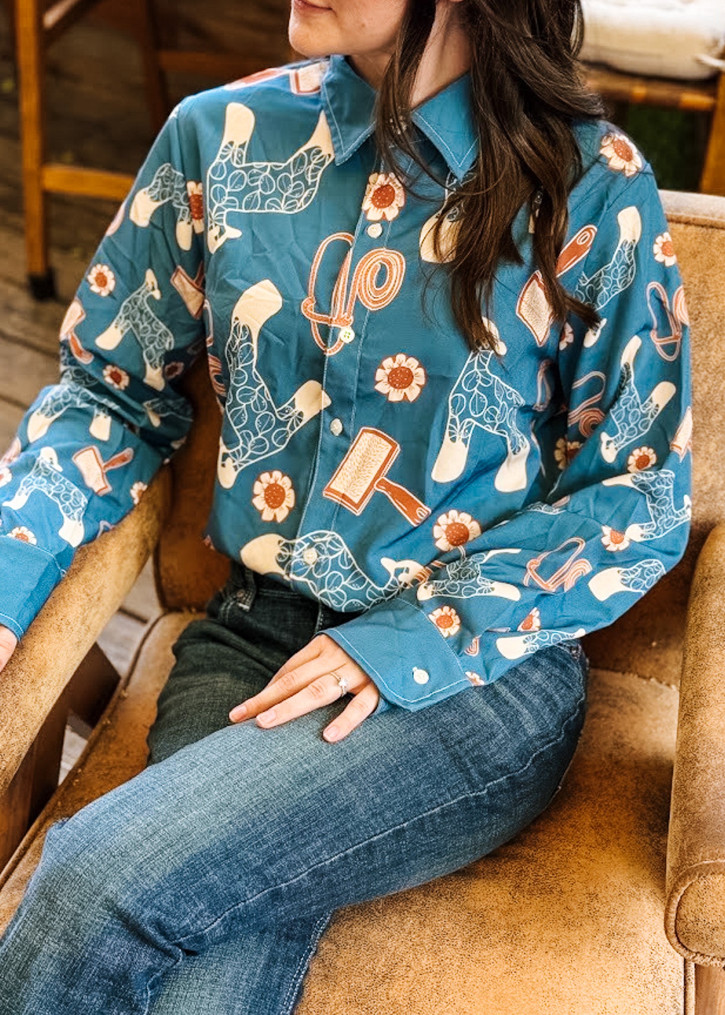 A woman wearing a long-sleeve button-up shirt with a western-style print featuring a teal background, white cowboy boots, brown and orange details, and blue jeans, seated on a tan armchair.