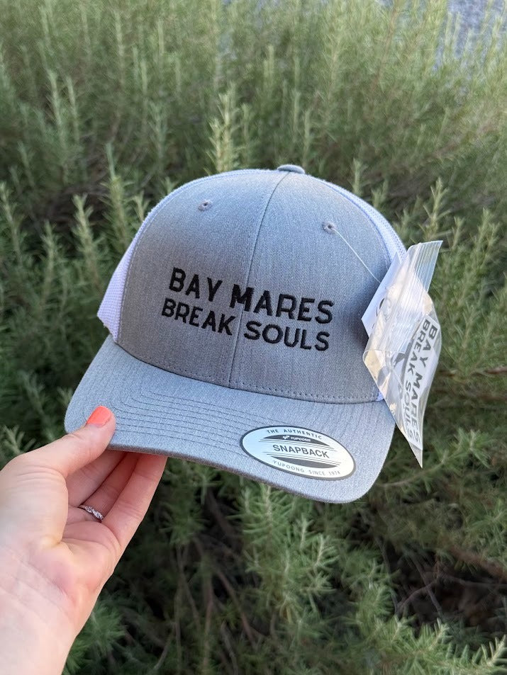 A light gray Snapback cap with black embroidered text reading 'BAY MARES BREAK SOULS' held by a hand against a background of green shrubs. The cap has a white Snapback snap closure and a hangtag attached.