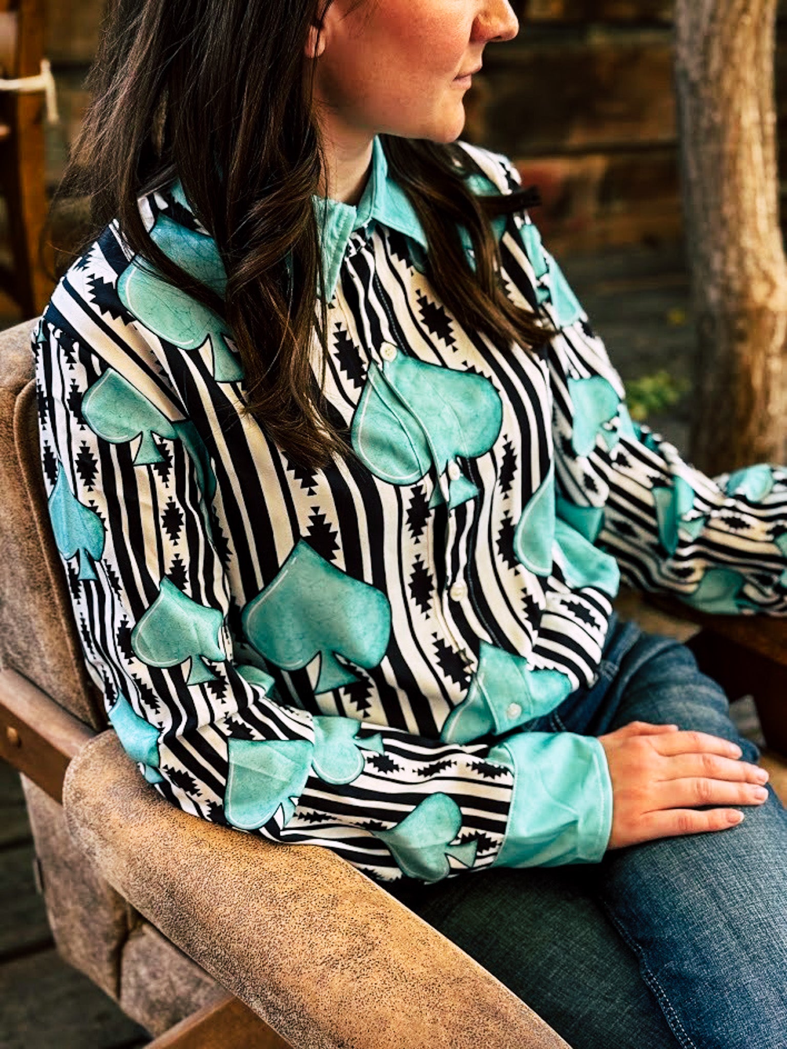 A woman wearing a long-sleeve button-up shirt with a black and white zebra stripe pattern and large turquoise spade-shaped appliques, seated in a rustic wooden chair.