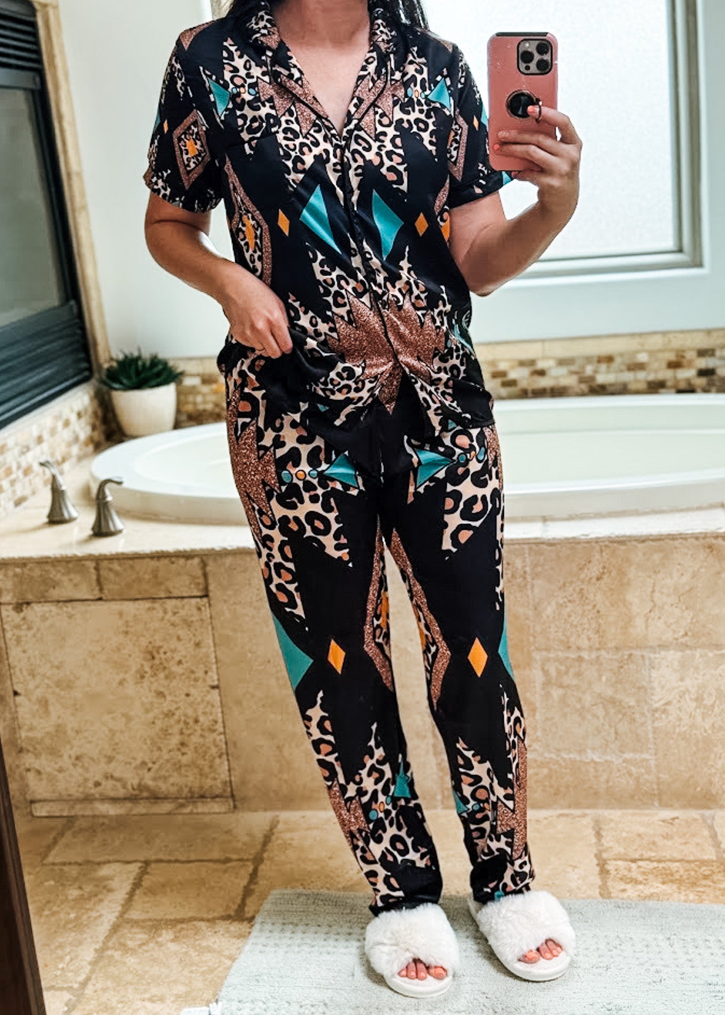 A woman takes a mirror selfie in a bathroom wearing a matching short-sleeve button-up pajama top and pants set. The set features a black base with a multicolor print combining leopard spots, geometric diamond shapes, and teal accents.