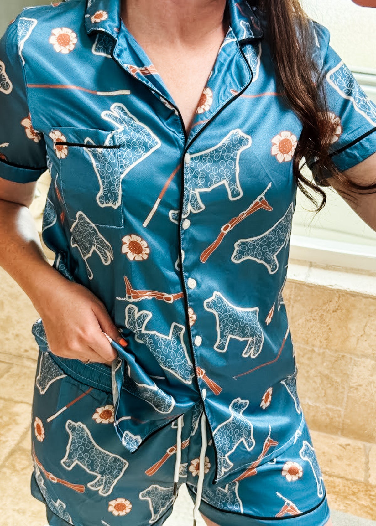 A person wearing a matching short-sleeve button-front pajama top and shorts set in teal blue with an allover print of white dogs, orange flowers, and orange-and-white rifles. The photo is cropped from the neck to the thighs, showing the pajama set in a real indoor setting with tiled walls and floor.