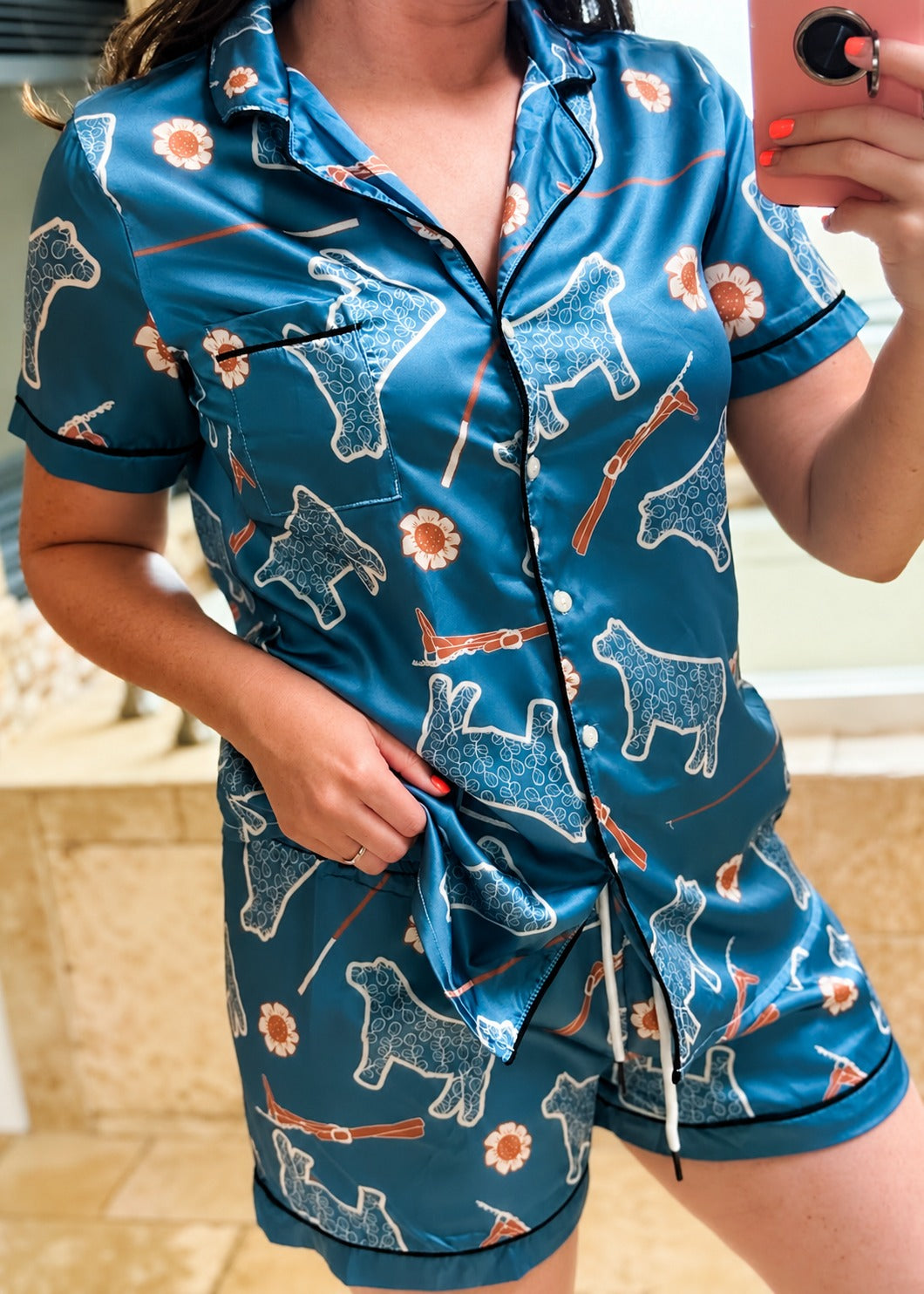 A woman wearing a teal blue satin pajama set with a dog print, taking a mirror selfie. The set includes a short-sleeve button-up shirt and matching shorts with black piping.