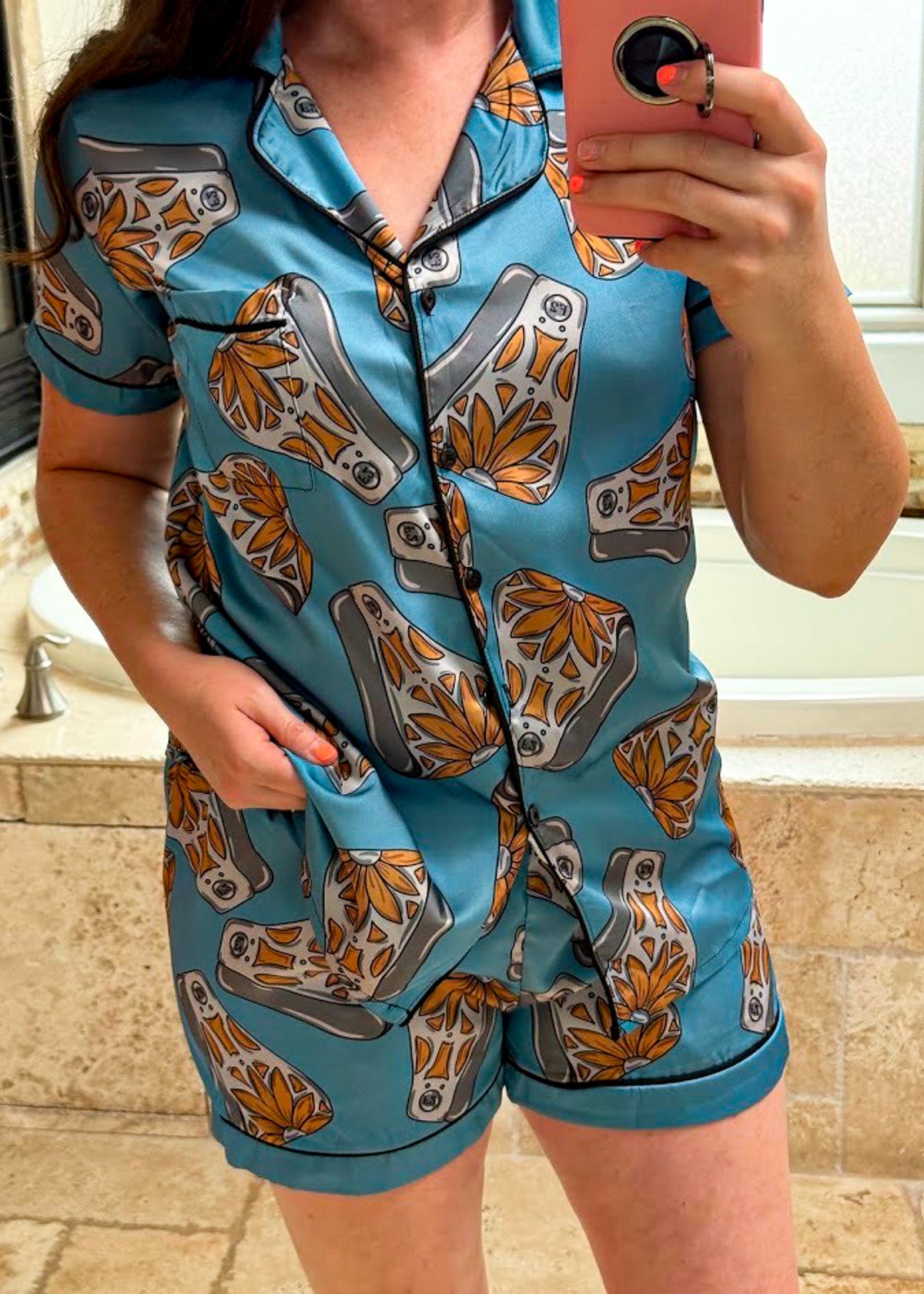 A person taking a mirror selfie in a bathroom wearing a short-sleeve pajama romper with a blue base and a pattern of gray and orange toilet paper rolls.