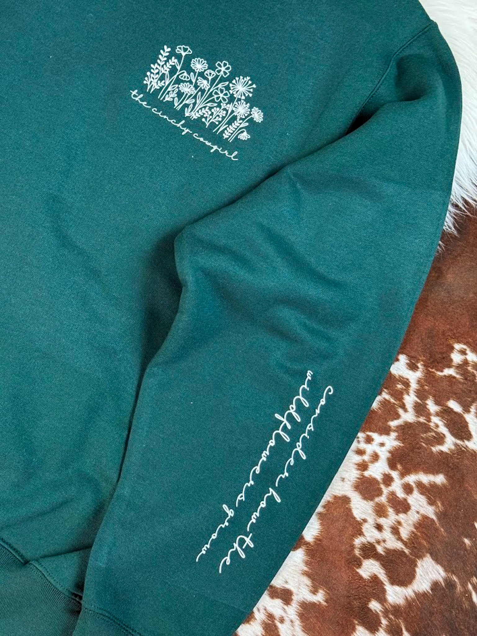 A close-up of a dark teal green sweatshirt featuring white embroidered graphics, including a floral design on the chest and cursive text on the sleeve, styled on a brown and white cowhide rug.