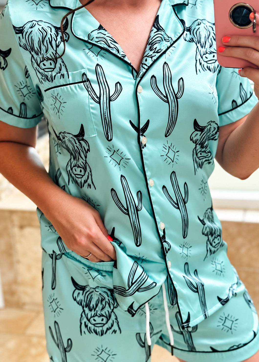 A person wearing a mint green short-sleeve pajama set with a black cactus and Highland cow print, taking a mirror selfie.