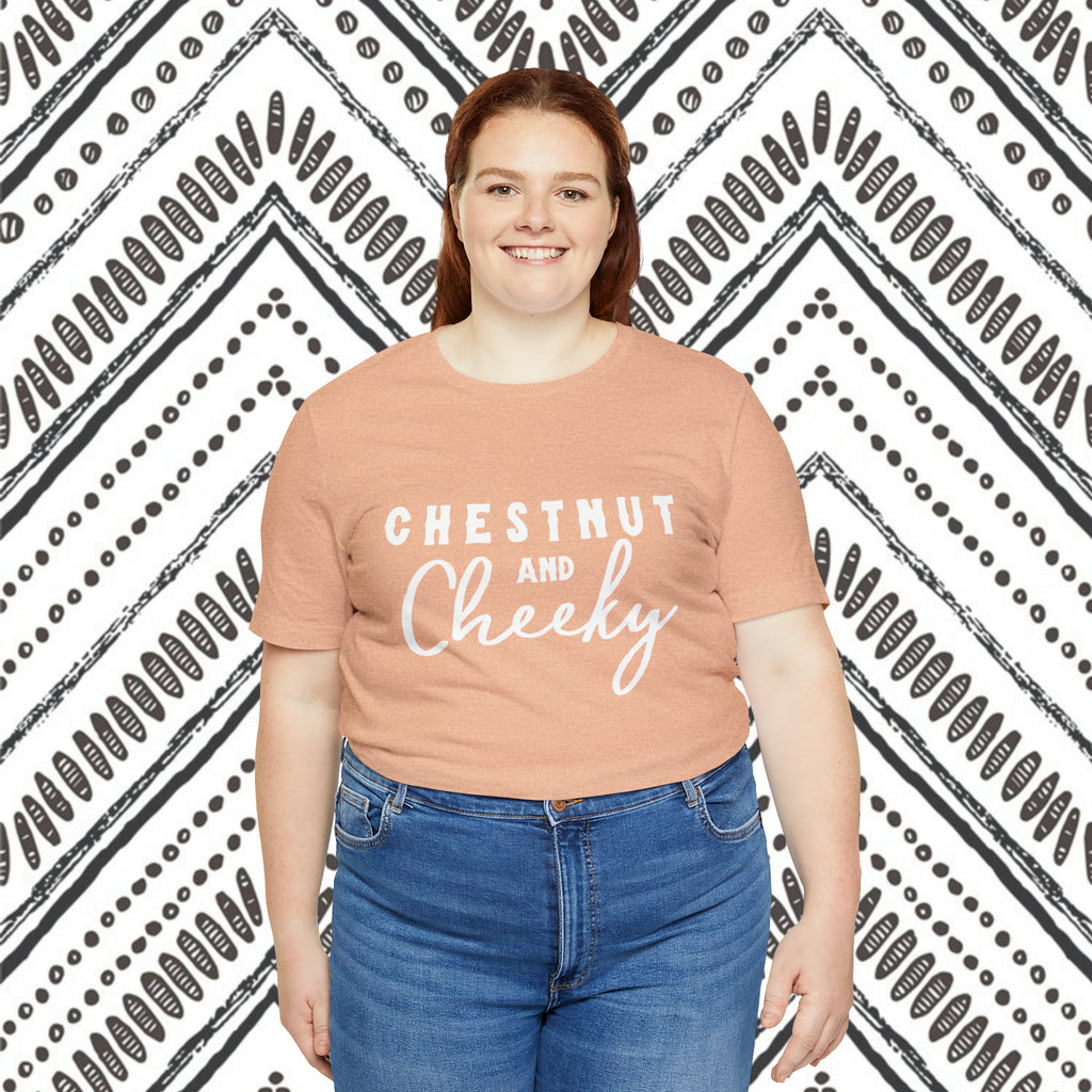 Peach plus size short sleeve with white lettering reading "Chestnut and Cheeky". Plus size model wears this against a black and white southwestern design background.