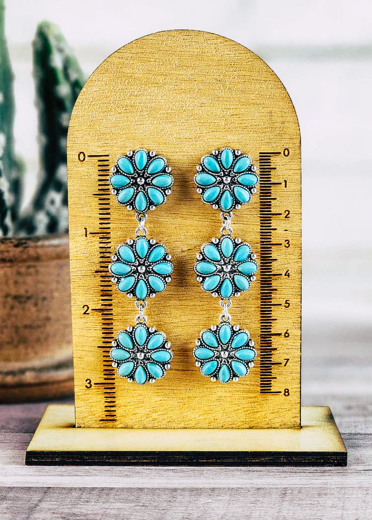 Western cowgirl earring with turquoise-like stone in floral concho. One concho acts as the stud with two of the same conchos dangling. Hangs about 3" on the measuring block. 