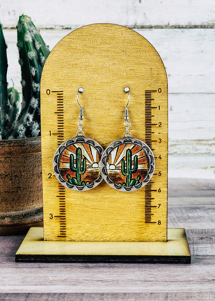 Desert sunset scenery with a setting sun decoration the sky and a cactus up front leather earrings with decorative embossments around the scene. Earrings hang about 2.5 inches. 