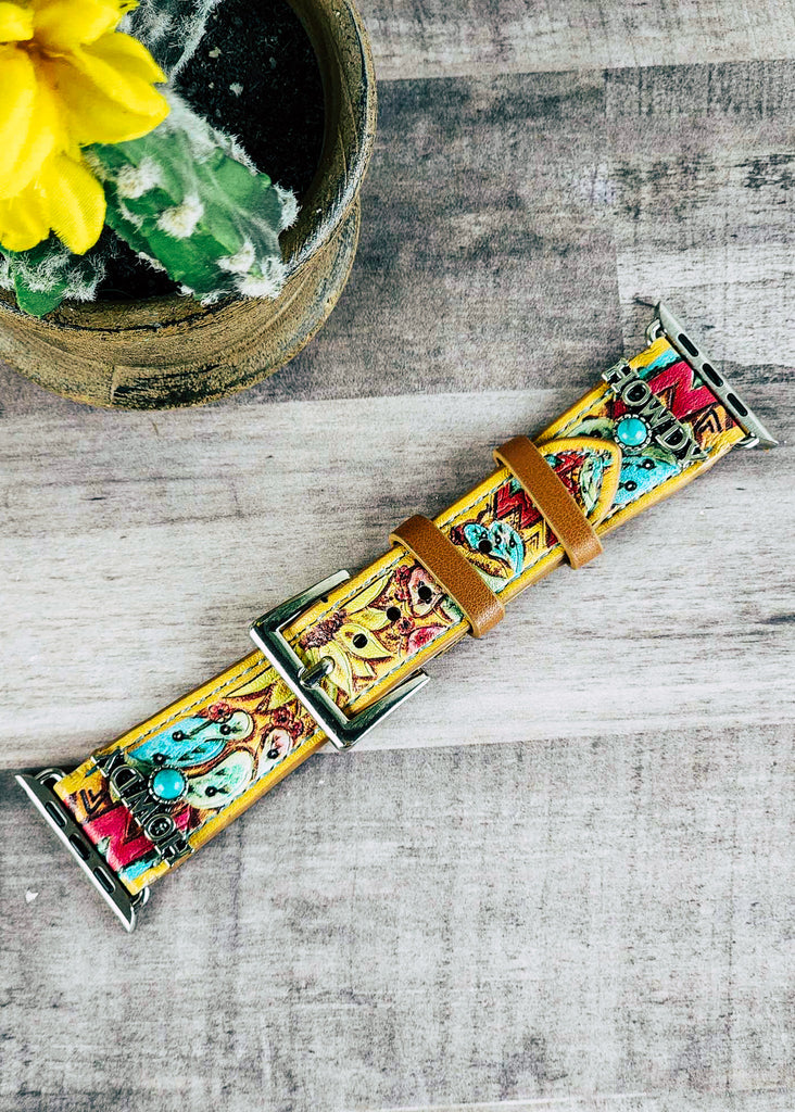 Yellow apple watch band with floral tooling painted. Watch band has silver accent reading "Howdy" with a turquoise colored stone. Band lays on wooden table. 