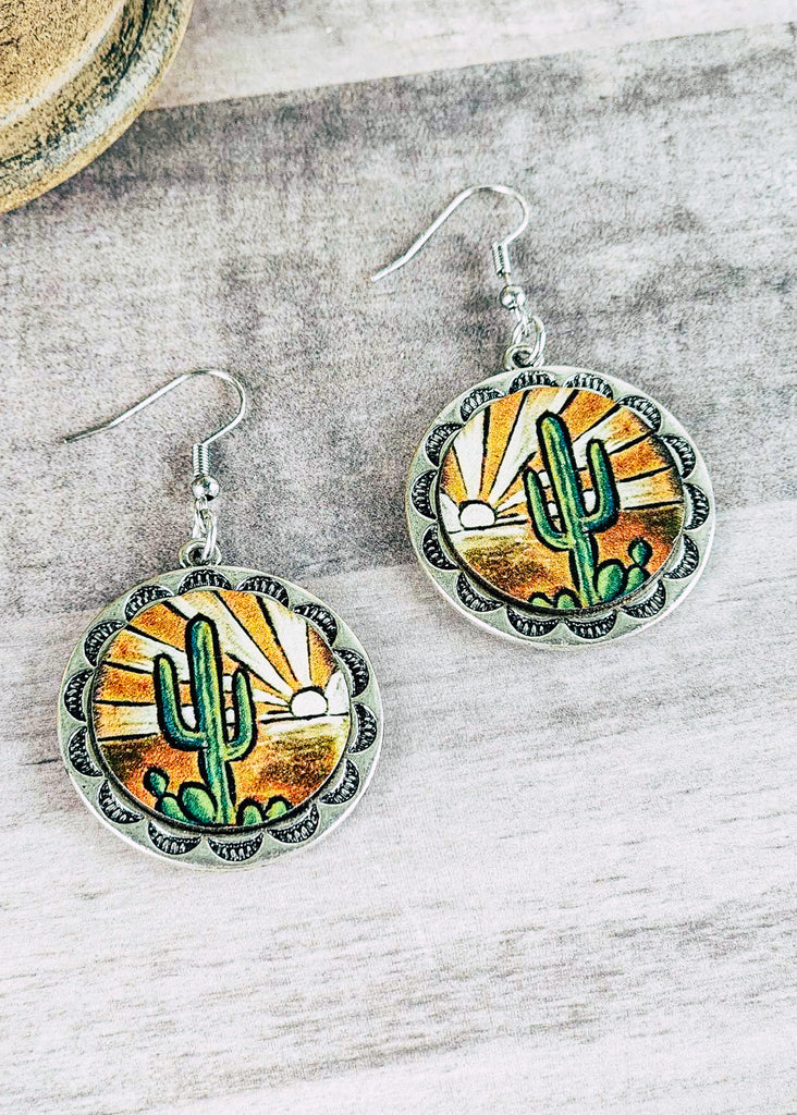 Desert sunset scenery with a setting sun decoration the sky and a cactus up front leather earrings  with decorative embossments around the scene. 