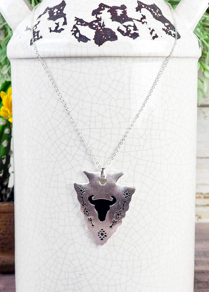 Southwestern cowgirl necklace with an arrowhead pendant. On the pendant is a stamped steer skull and tribal design with arrows. Pendant is connected to a chain necklace and is hanging on a rustic milk can. 
