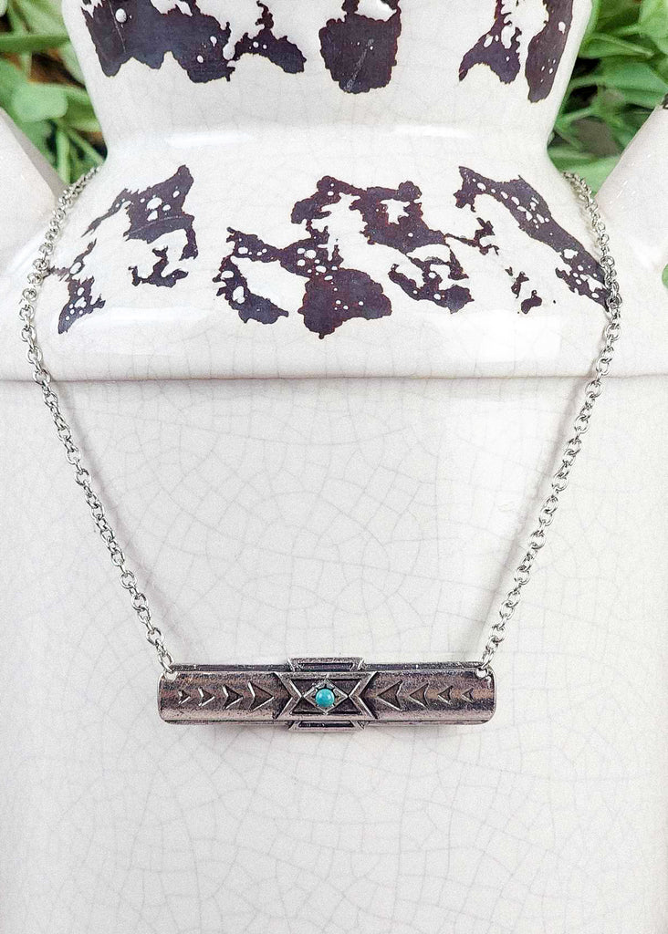 Southwestern cowgirl necklace with a tribal bar necklace pendant with a turquoise-like stone in the middle. Chain holding the necklace. Hanging on a rustic milk jug. 