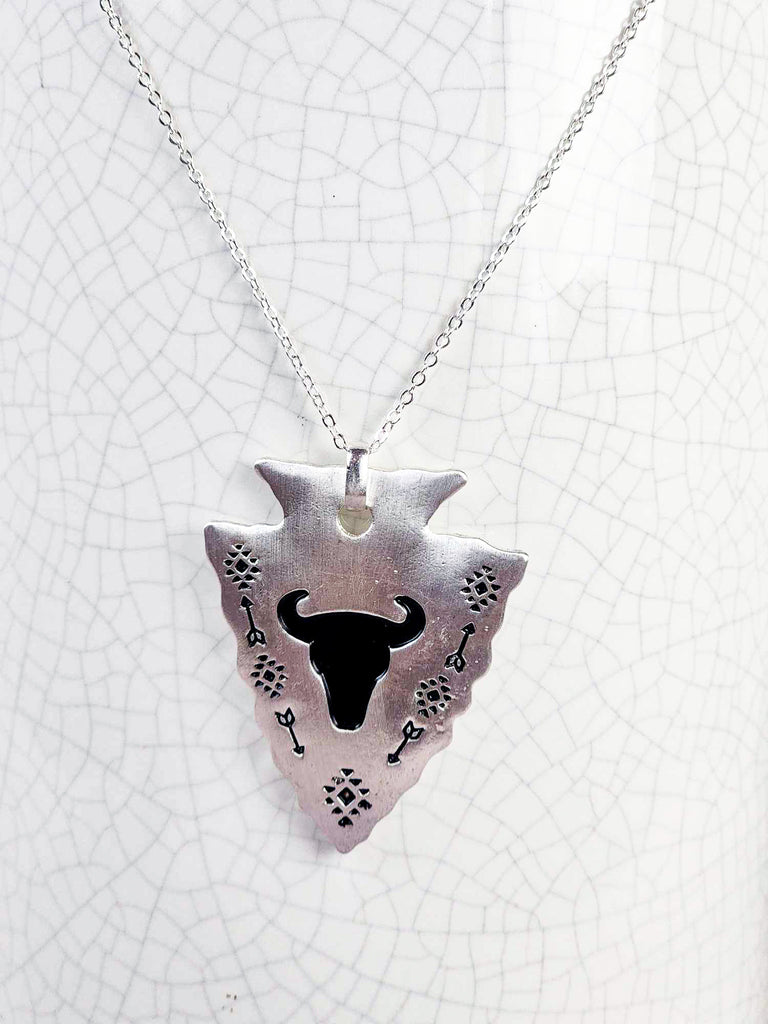 Southwestern cowgirl necklace with an arrowhead pendant. On the pendant is a stamped steer skull and tribal design with arrows. Pendant is connected to a chain necklace and is hanging on a rustic milk jug. 