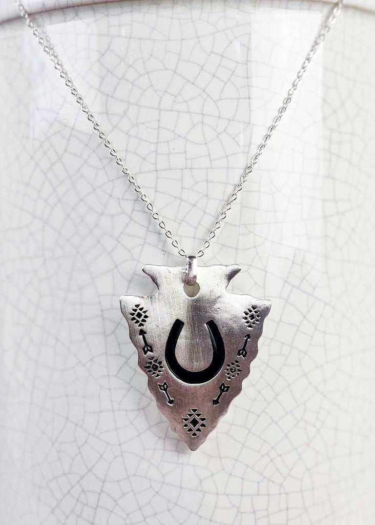 Southwestern cowgirl necklace with an arrowhead pendant. On the pendant is a stamped horseshoe and tribal design with arrows. Pendant is connected to a chain necklace and is hanging on a rustic milk jug. 