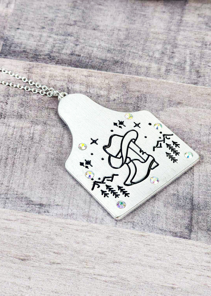 Cow tag with engraved boot and cowboy hat design in the front of a  mountain scape. Sparkle jewels circle the edges of the cow tag. Pendant is on the chain. 