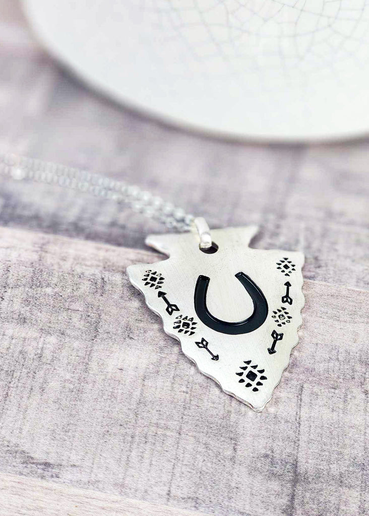Southwestern cowgirl necklace with an arrowhead pendant. On the pendant is a stamped horseshoe and tribal design with arrows. Pendant is connected to a chain necklace and is laying on a wooden table.