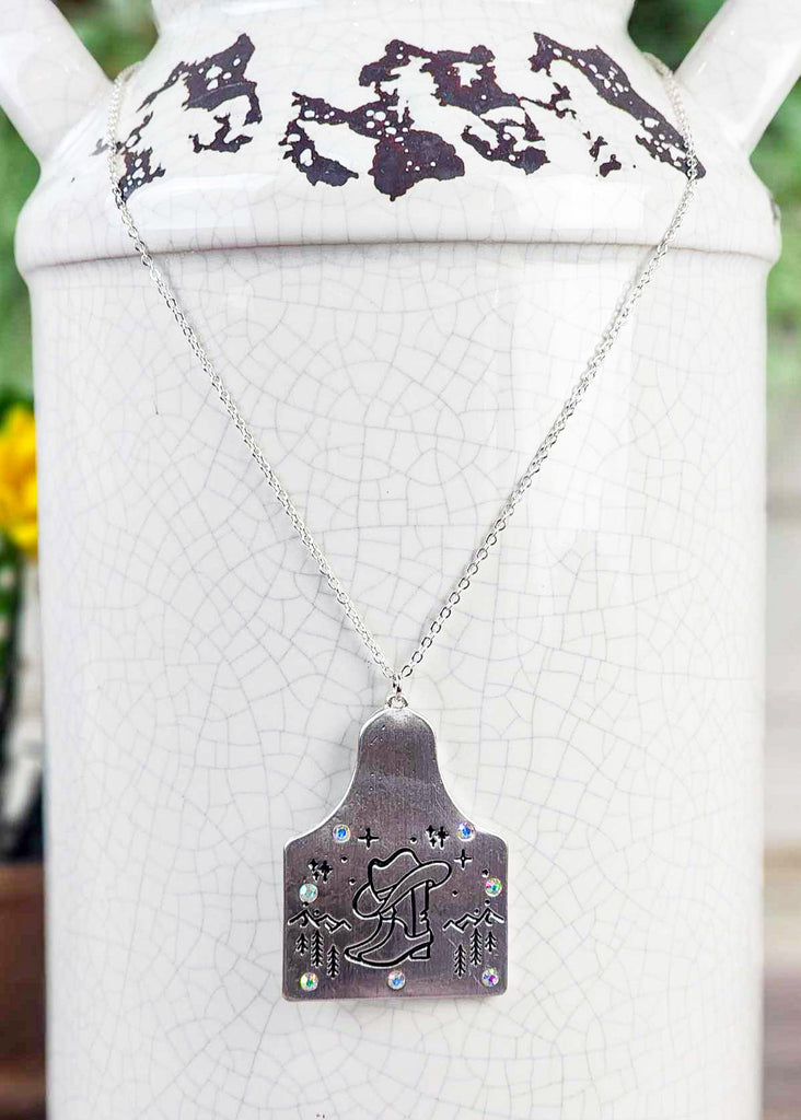 Cow tag with engraved boot and cowboy hat design in the front of a  mountain scape. Sparkle jewels circle the edges of the cow tag. Pendant is on the chain.  Necklace hangs around the rustic jar.