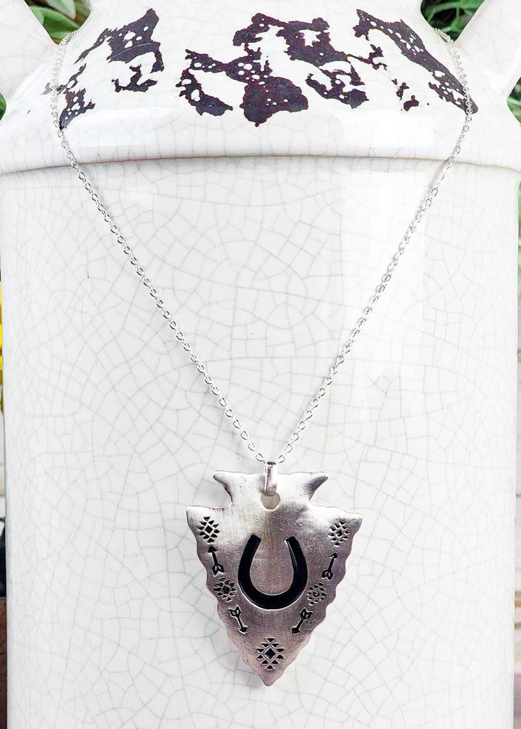Southwestern cowgirl necklace with an arrowhead pendant. On the pendant is a stamped horseshoe and tribal design with arrows. Pendant is connected to a chain necklace and is hanging on a rustic milk jug. 