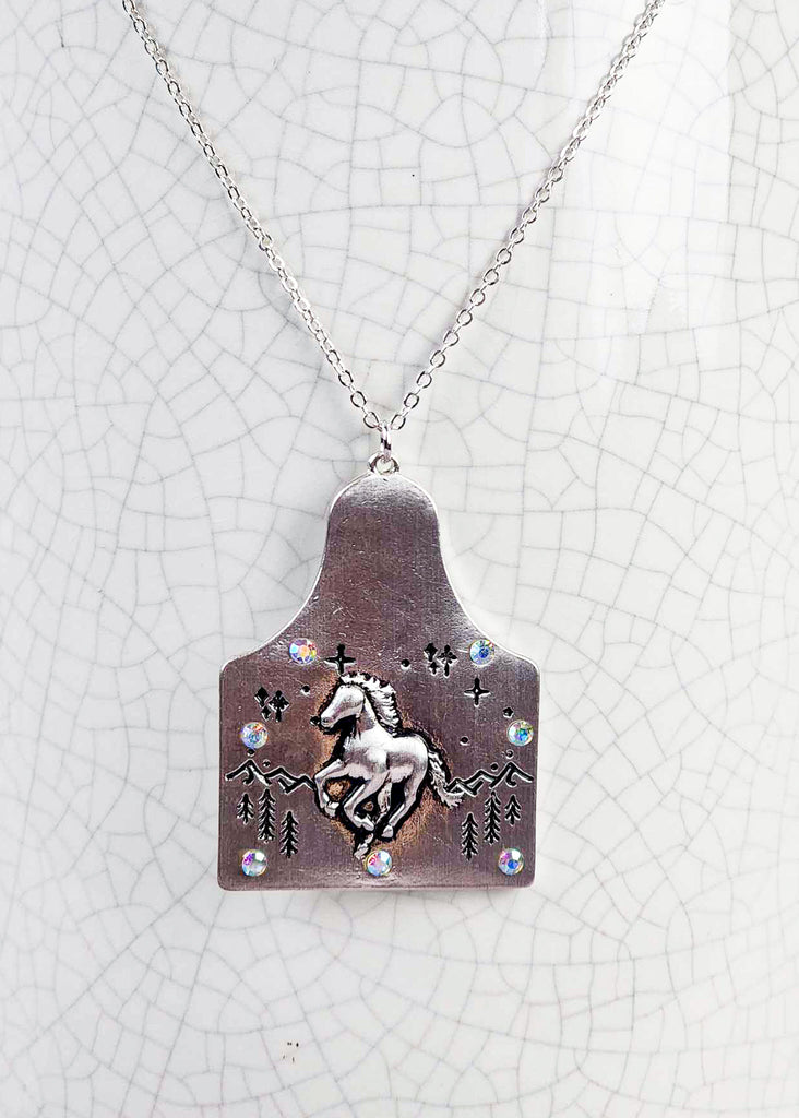 Cow tag with engraved wild running horse design in the front of a  mountain scape. Sparkle jewels circle the edges of the cow tag. Pendant is on the chain. 