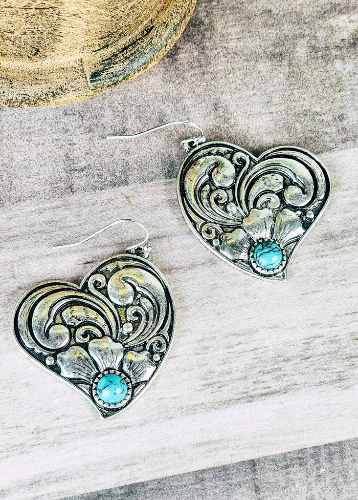 Western cowgirl heart earrings antique silver floral design and a turquoise-like stone acting as the middle of flower. Laying on wooden table.