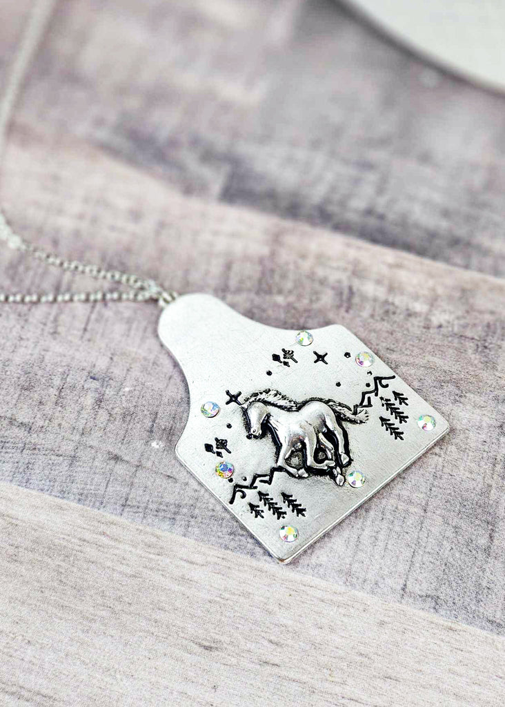 Cow tag with engraved wild running horse design in the front of a  mountain scape. Sparkle jewels circle the edges of the cow tag. Pendant is on the chain. 