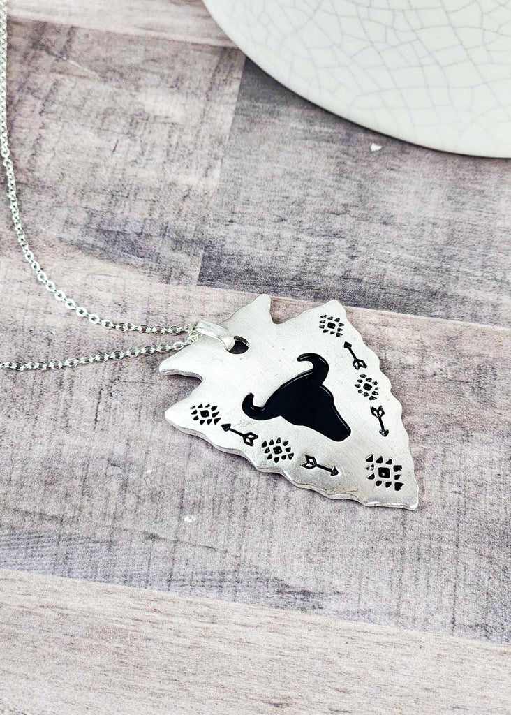 Southwestern cowgirl necklace with an arrowhead pendant. On the pendant is a stamped steer skull and tribal design with arrows. Pendant is connected to a chain necklace and is laying on a wooden table.