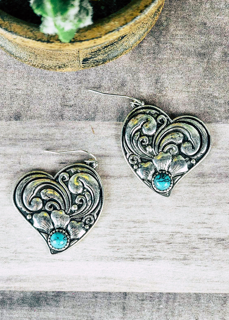 Western cowgirl heart earring antique silver floral design and a turquoise-like stone acting as the middle of flower. Laying on wooden table.