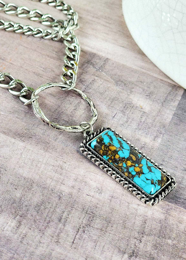 Edgy cowgirl necklace with two layers of 'dog chain' attached to a turquoise like stone by an O-ring. Accents of antique silver are encasing the turquoise-like stone. Zoomed into the stone hints of gold specks and turquoise blue are visible. 