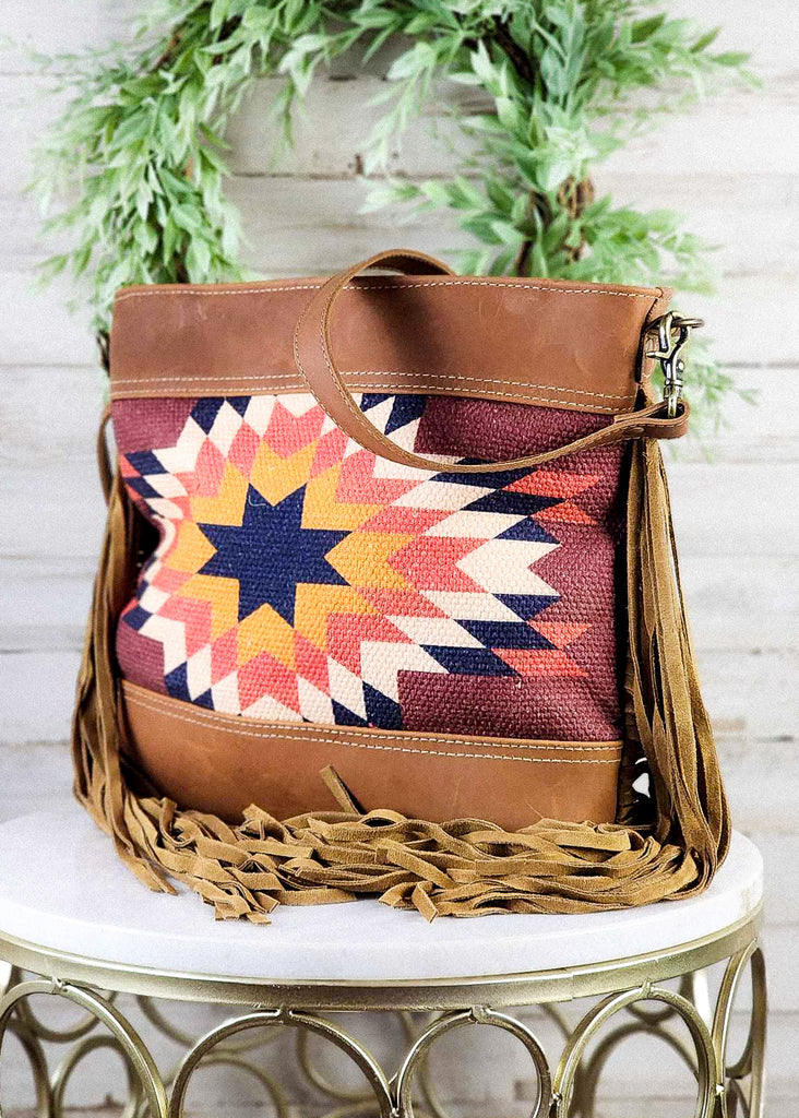 Angled view of the purse. Fringe is wrapped to center of the purse with should strap draped over teh top of the purse. Leather panels and woven rug is shown going to the side seam . 