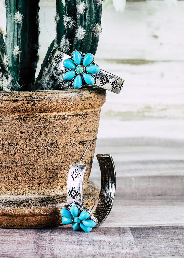 Cowgirl hoop stud earring with floral turquoise like stone design. Antique silver stamped hoops. Laying next to pot on wood counter and in pot next to cactus. 