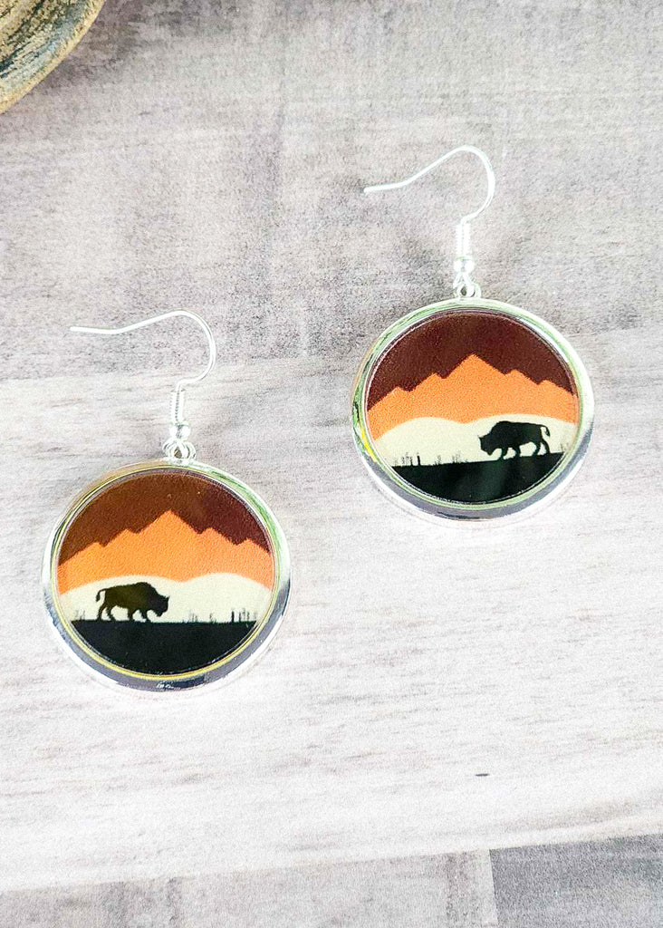 Southwestern buffalo scenery earring with orange and reds matching a sunset sky on a wooden table. 