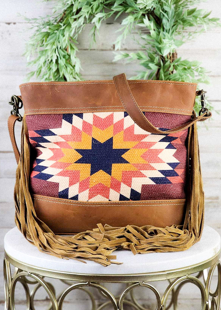 Western style fringe purse sitting on a marble stool. The fringe is brought to the middle of the bag with the shoulder strap draped over the top of the purse. Blue, maroon, blue, yellow, and white are the main colors of the Sedona star.