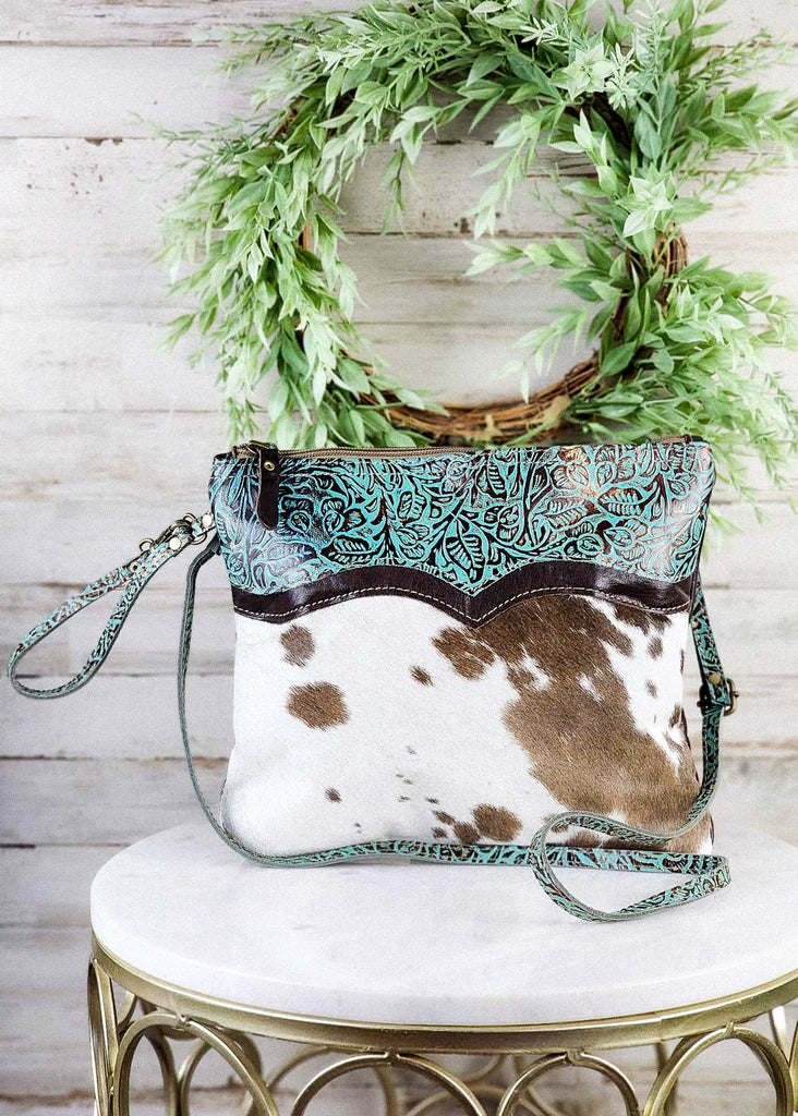 Front View. Matching teal tooled leather accent, shoulder strap, and wristlet are visible. White and brown colored cowhide. Purse sitting on marble stool. 