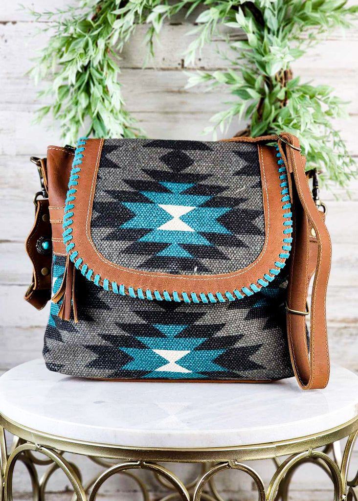 Cowgirl starfire purse with black, grey, and turquoise detailing. Leather accents around the woven material with matching straps. Teal hand-stitch detail around top flap of purse. 