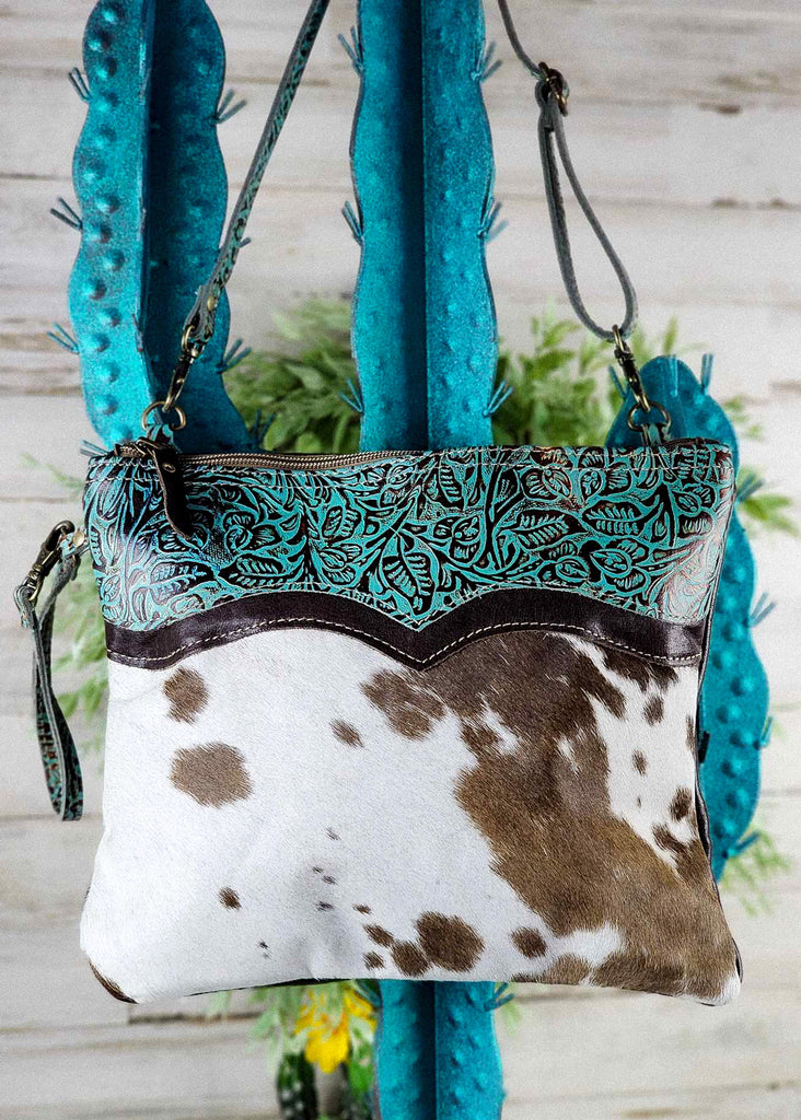 Close Up Hanging View. Western Cowhide and tooled leather with teal accent handbag hanging from a blue cactus. 