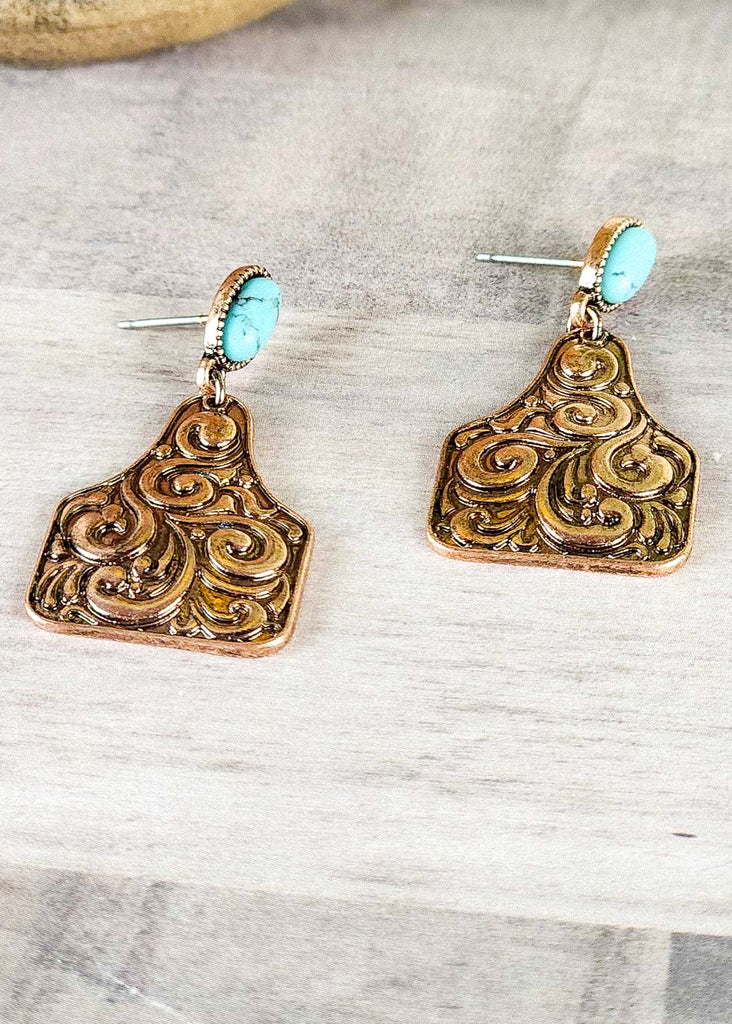 Cowgirl earring with metal engraved cow tag with copper finish. Turquoise like stone facing the side showing the length of the stud.  