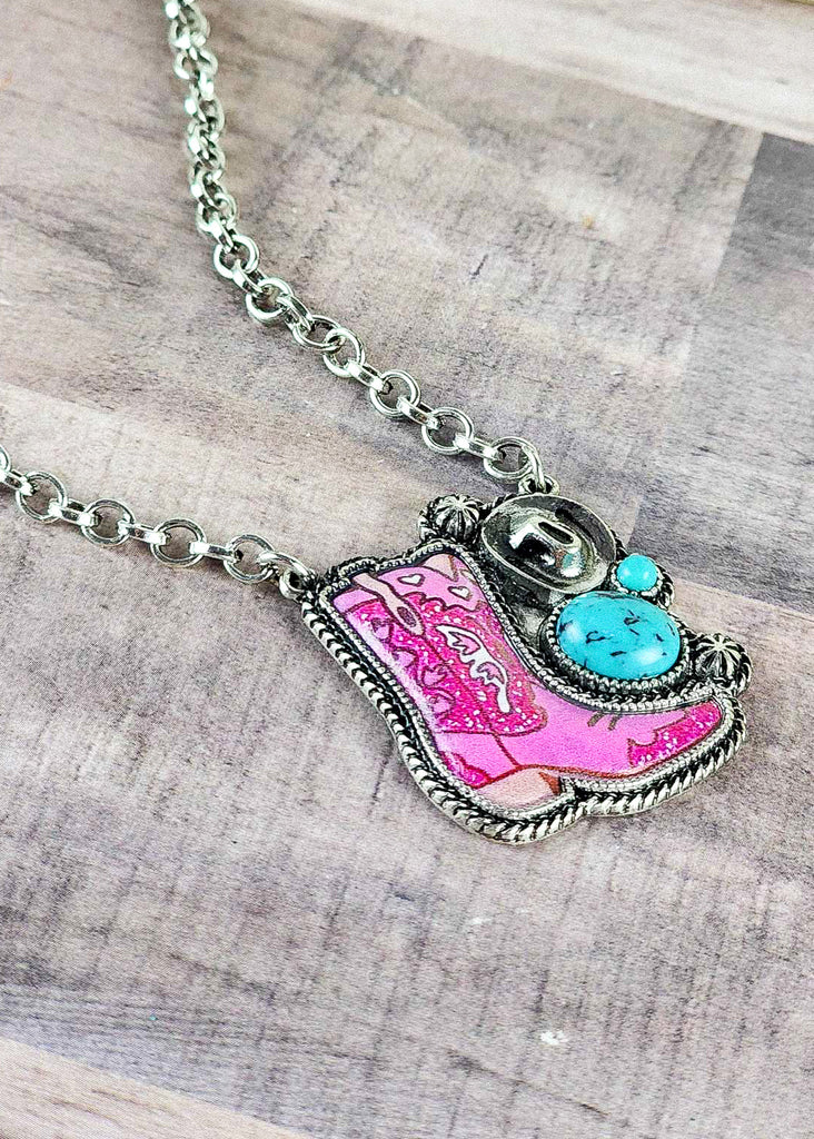 Cowgirl  necklace with bright pink epoxy boot design with two turquoise like stones and antique silver designs including a cowboy hat and conchos. 