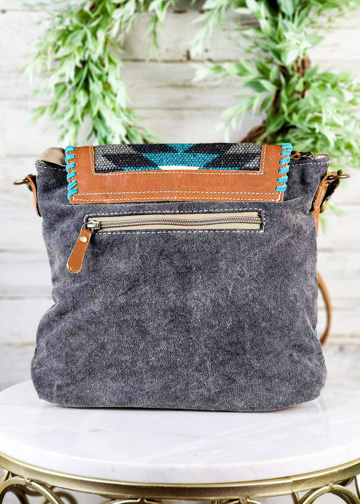 Backside of purse. Dark grey canvas with zippered pocket. Leather accent from top flap, hand stitching, and some of the tribal pattern visible. 