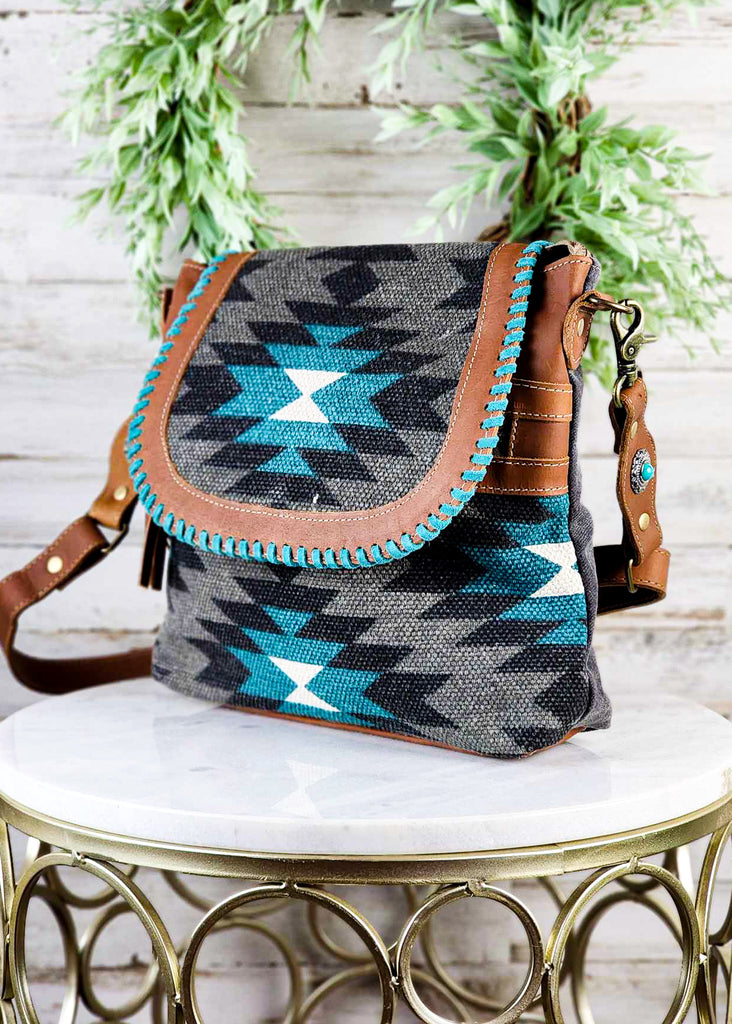 Angled view of this western purse. Canvas woven material with a tribal pattern and leather accents. Turquoise Hand stitching can be seen.  Adjustable shoulder strap with turquoise concho on strap. 