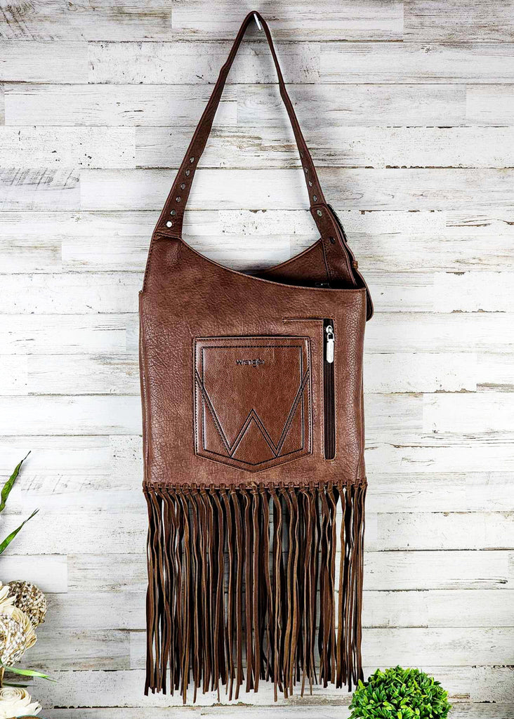 Coffee wrangle hobo with fringe handing up against a wooden wall. Silver studded adjustable shoulder strap. Zippered pocket seen.