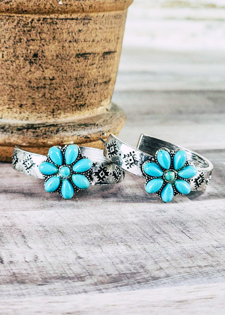 Cowgirl hoop stud earring with floral turquoise like stone design. Antique silver aztec design stamped hoops. Laying next to pot on wood counter. 