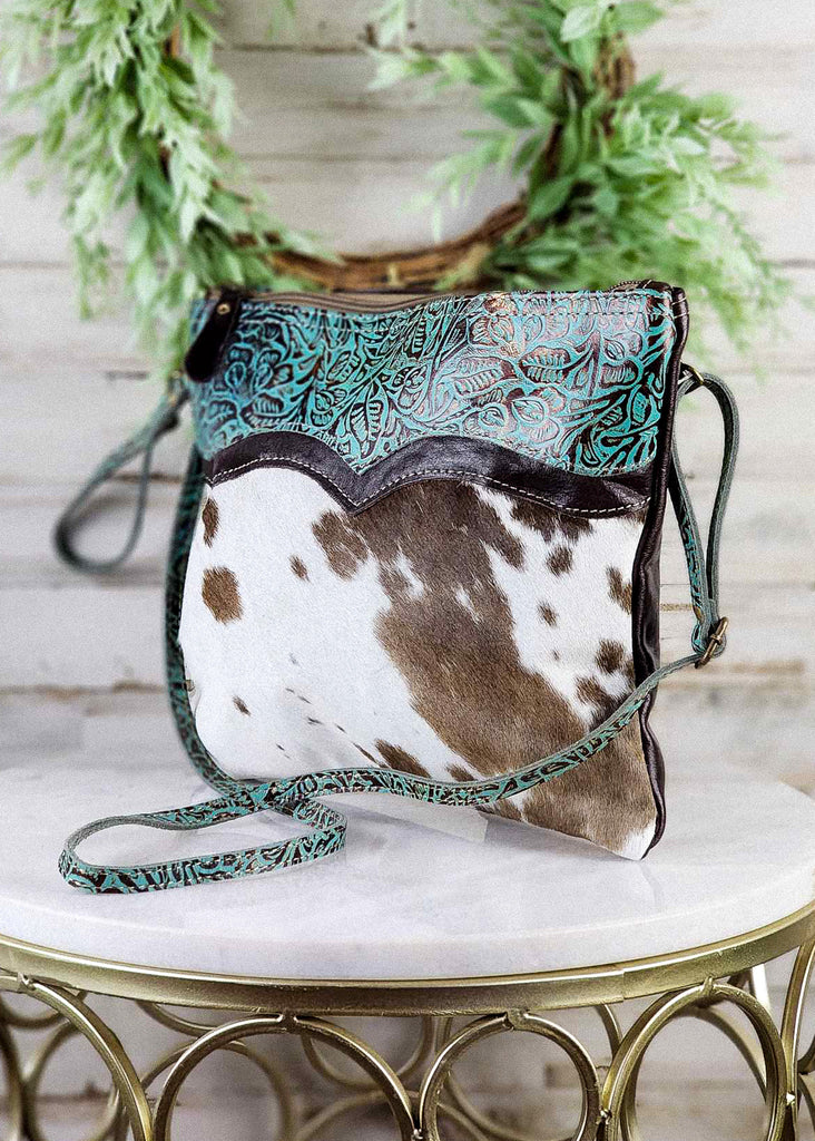 Side Angle. Tooled leather and cowhide shown to side seam. Matching tooled leather shoulder strap and wristlet seen. Shoulder strap is to the front of the purse. 