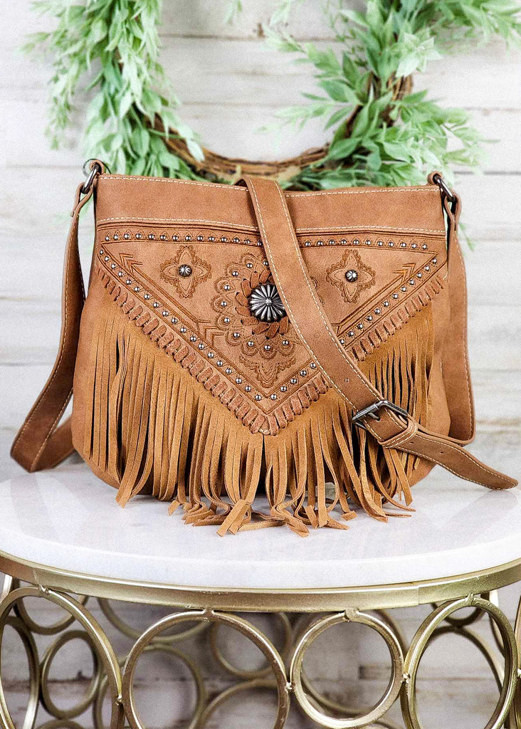 Western concealed carry fringe bag with antique silver concho and floral rough out. Matching leather fringe and adjustable shoulder straps. Whip stitch and studs follow fringe and top of purse. Bag sits on marble stool. 