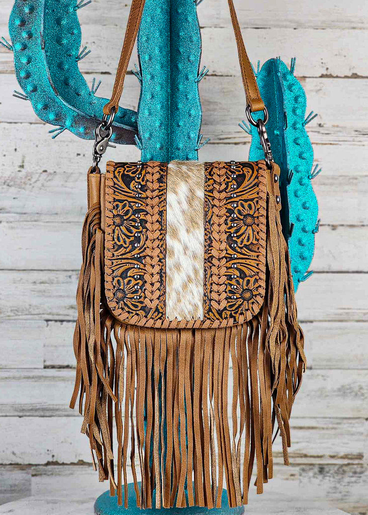 Western cowgirl hand bag with floral tooling and cowhide flap. Flap additional details include leather whip stitching and silver studs. Fringe hangs at bottom and sides of bag. Adjustable and removable shoulder strap are visible. Purse hangs from blue metal cactus rack. 
