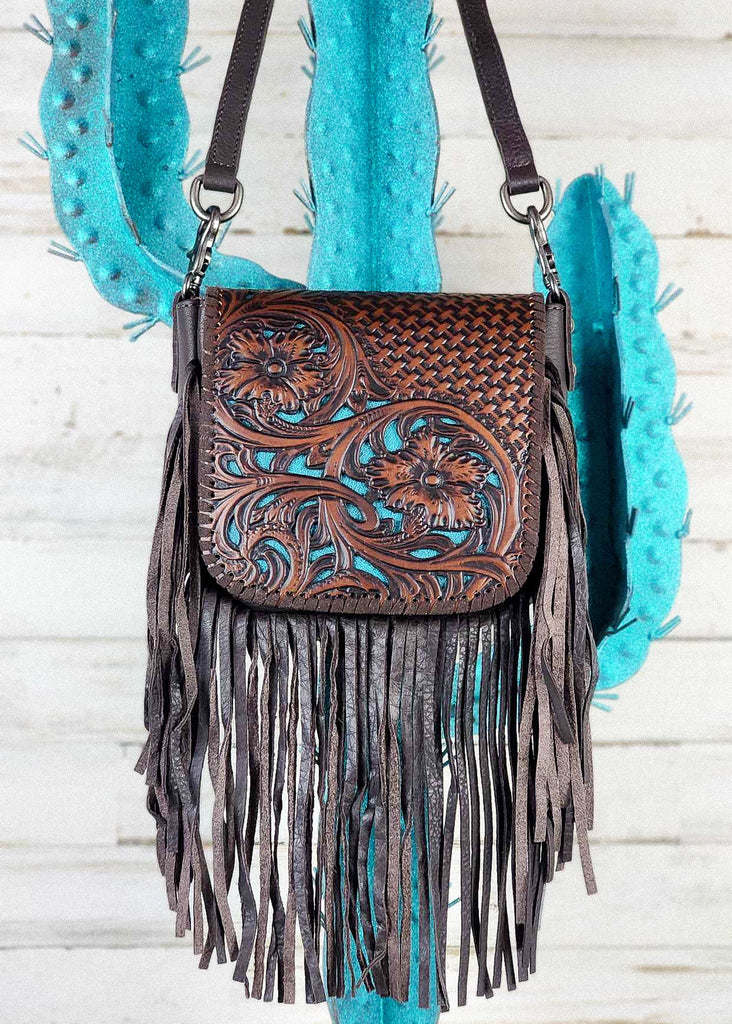 Western cowgirl purse with coffee floral and basket weaved tooling and matching leather whip stitch. Underneath floral tooling is a turquoise base to make floral design pop. Removable shoulder strap clasp are visible. Purse is hanging from a blue cactus metal rack. 