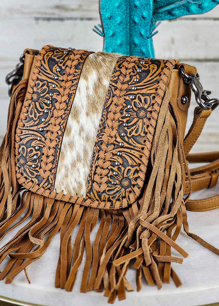 Western cowgirl hand bag with floral tooling and cowhide flap. Flap additional details include leather whip stitching and silver studs. Fringe hangs at bottom and sides of bag. Adjustable and removable shoulder strap are visible. 