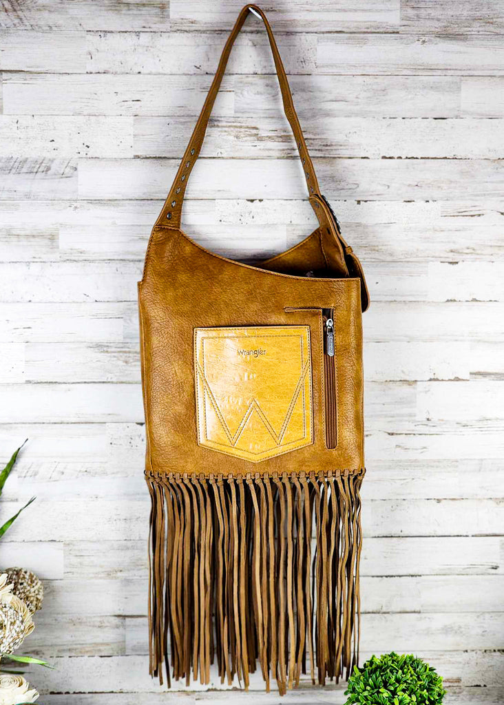 Brown wrangle hobo with fringe handing up against a wooden wall. Silver studded adjustable shoulder strap. Zippered pocket seen. 