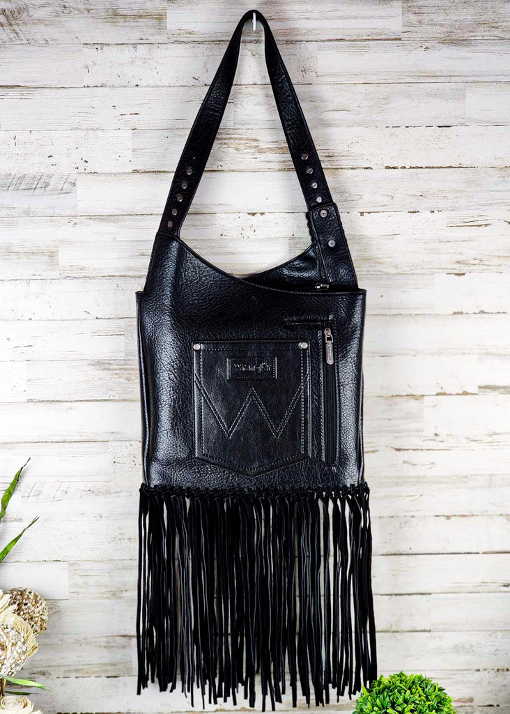 Black wrangle hobo with fringe handing up against a wooden wall. Silver studded adjustable shoulder strap. Zippered pocket seen.